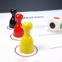 Drei Spielfiguren in Schwarz Rot und Gold, ein Wahlzettel und ein Würfel